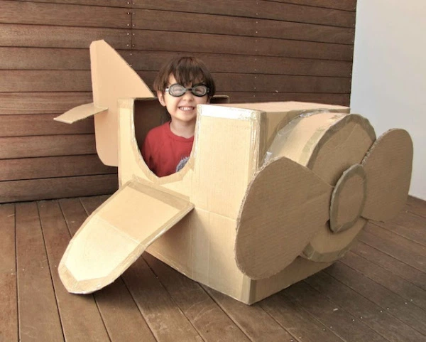 smiling little boy wearing aviation goggles sits in an airplane made of cardboard